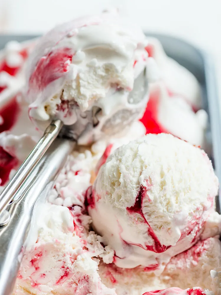 A scoop spoon scooping out some strawberries and cream ice cream from a tub