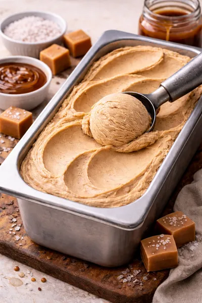 A metal tub of sea salted caramel gelato with a scooper resting on top all surrounded by chunks of caramel sprinkled with seal salt